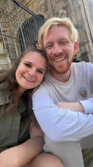 Mel and Scott smiling in a sunny selfie outside an old stone building