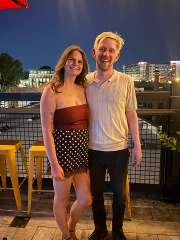 Mel and Scott standing together on a rooftop at night with city lights behind them