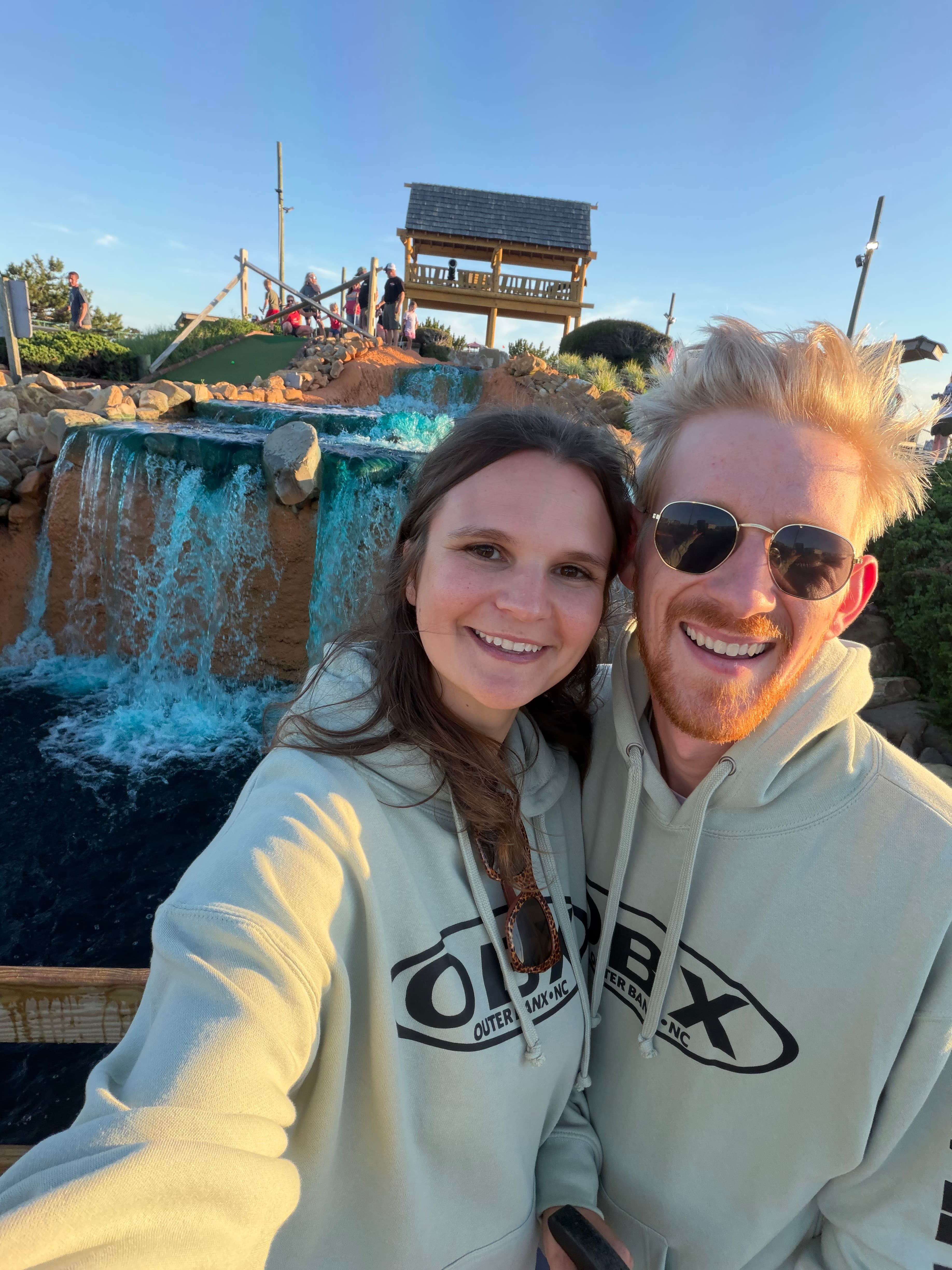 Mel and Scott taking a smiling selfie outdoors in matching Outer Banks sweatshirts